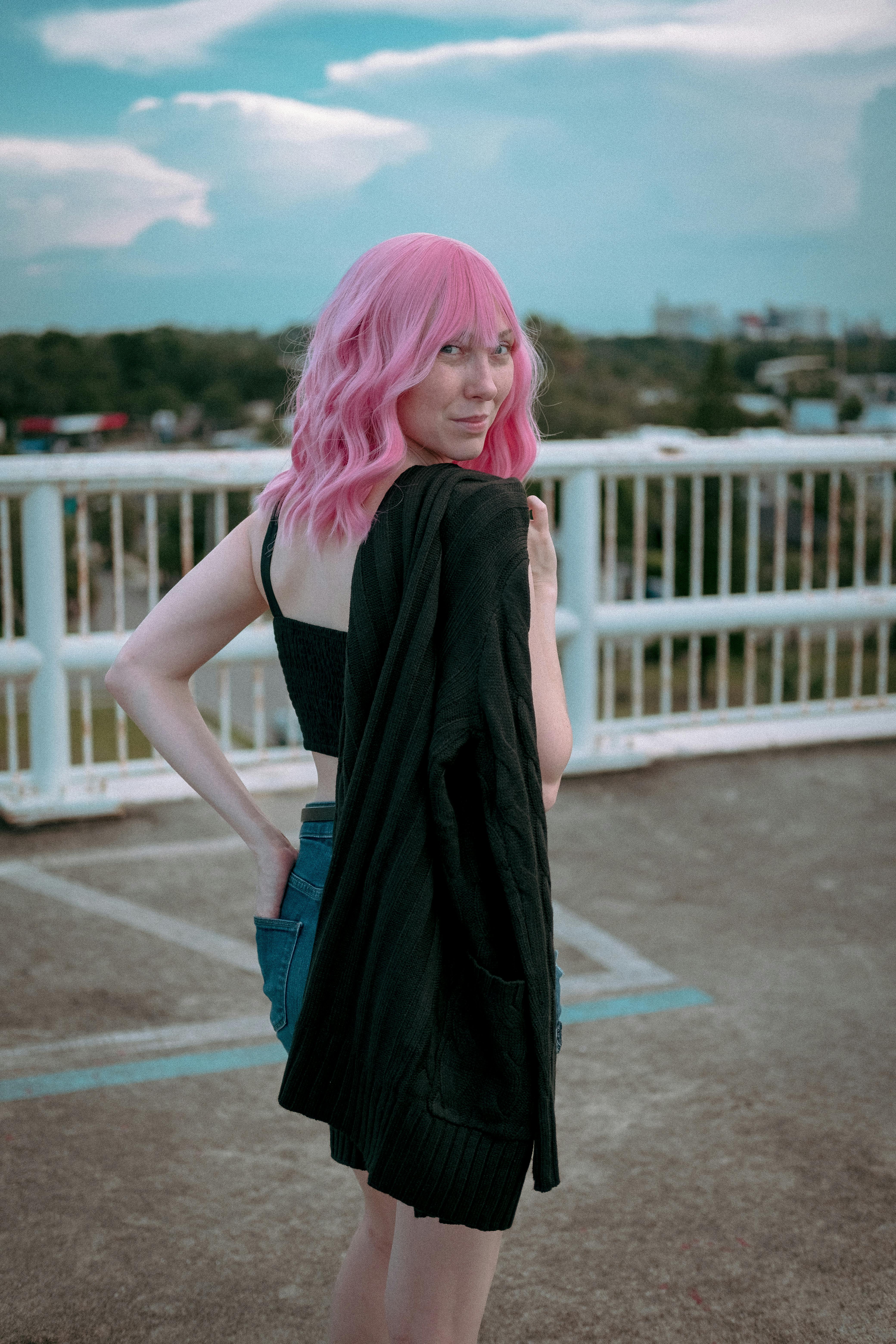 Free Stylish woman with vibrant pink hair posing outdoors, wearing a black top and denim shorts. Stock Photo