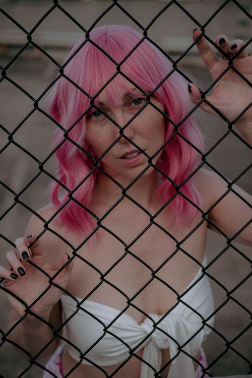 Free Woman with vibrant pink hair posing behind a chain link fence in an artistic outdoor photo. Stock Photo