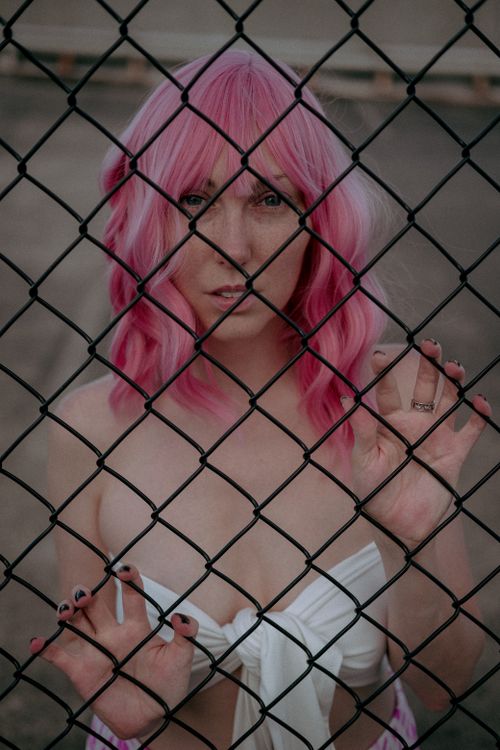 Free A woman with vibrant pink hair stands behind a chain link fence, expressing introspective mood. Stock Photo