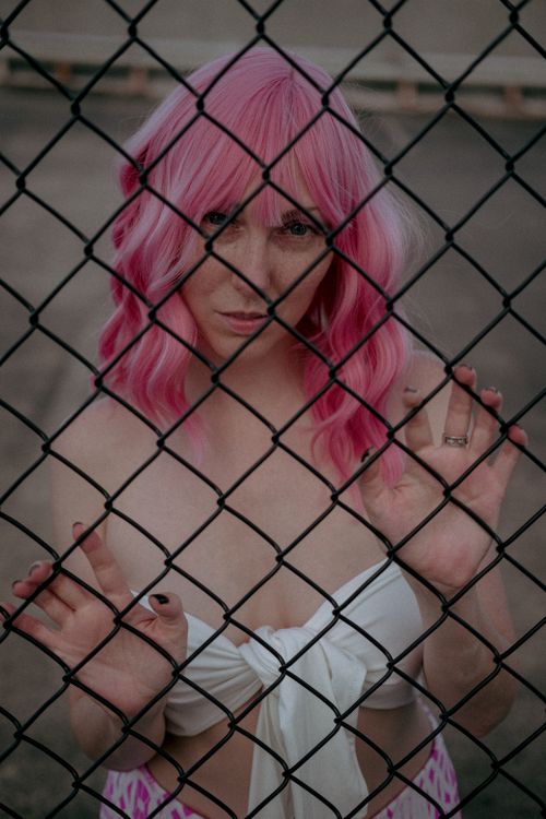 Free A woman with vibrant pink hair poses behind a chain link fence, wearing a white top. Stock Photo