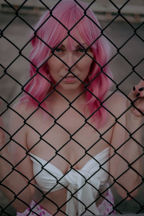 Free A woman with vibrant pink hair stands behind a chain link fence, wearing a white crop top. Stock Photo