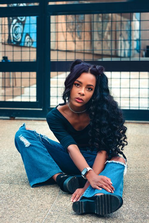 Free Young African American woman with curly hair sitting on street in casual attire, exuding modern style. Stock Photo