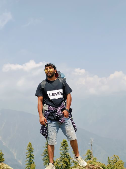 Free A man in casual attire poses on a mountain hike with a stunning backdrop of trees and sky. Stock Photo