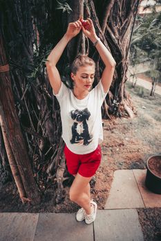 Fashionable woman wearing white shirt and red shorts posing by tree with arms raised.