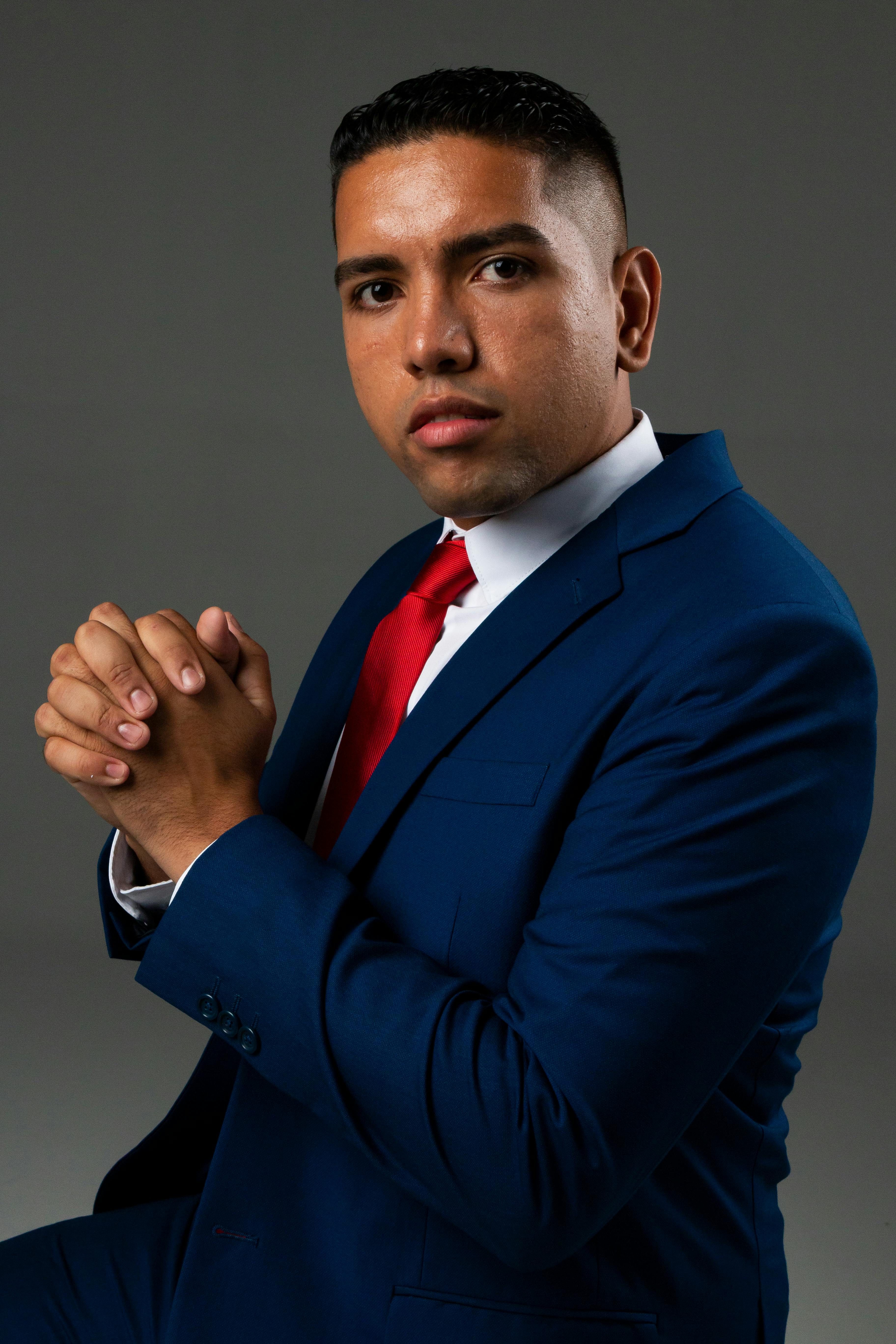 Professional studio portrait of a serious businessman in a blue suit with a red tie, hands clasped.
