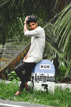 A stylish young man poses near a boundary marker amidst lush palm leaves.