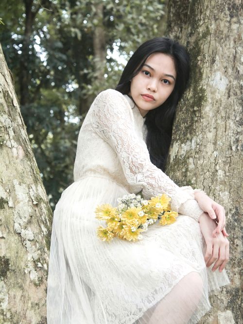 Free A young woman in a lace dress holds yellow flowers while seated against a tree outdoors. Stock Photo