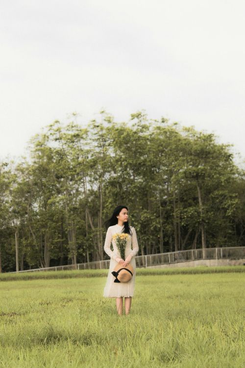 Free A woman in a dress stands in a lush field holding a hat, surrounded by trees. Stock Photo