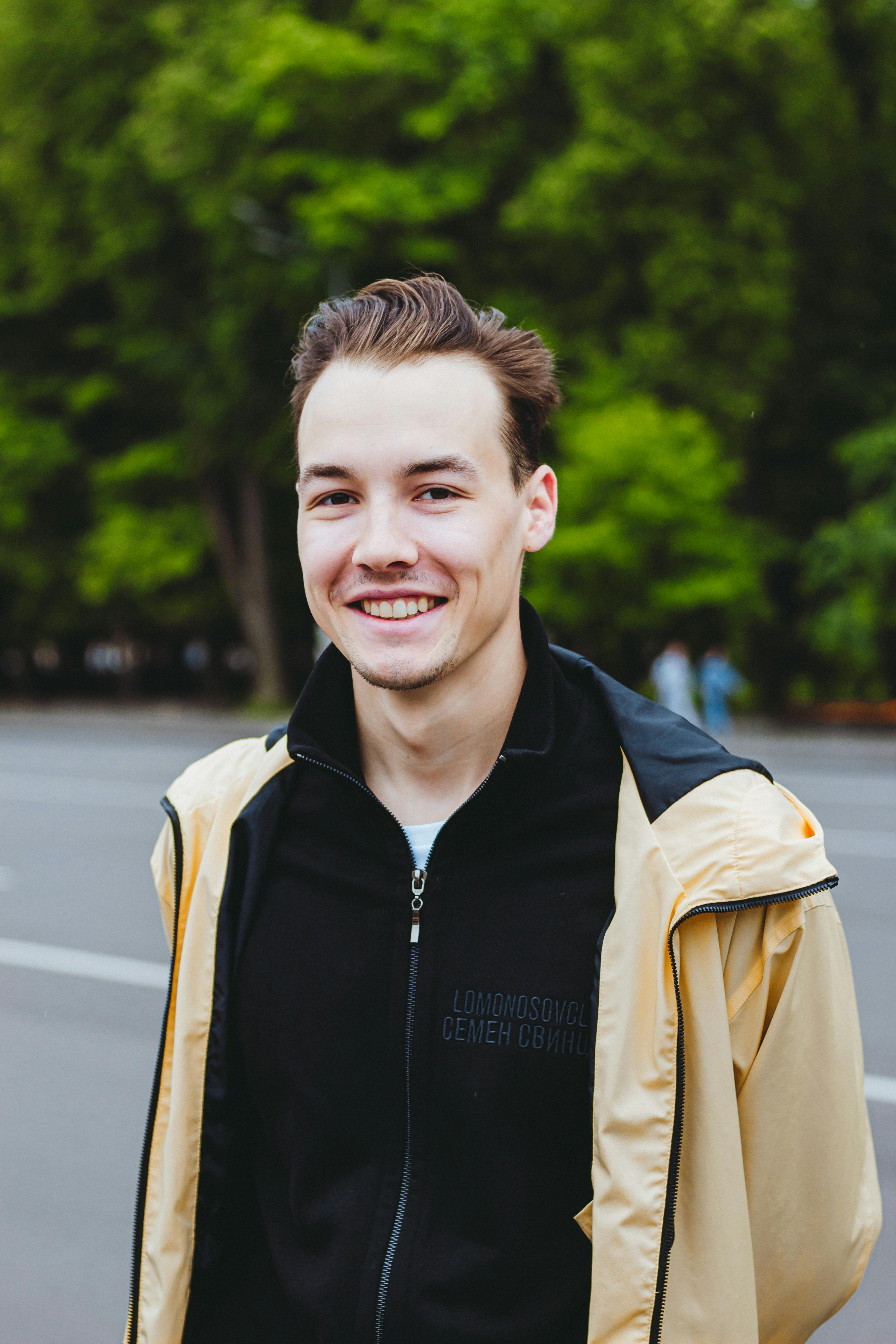 A smiling young man in casual attire poses in a lush urban park.