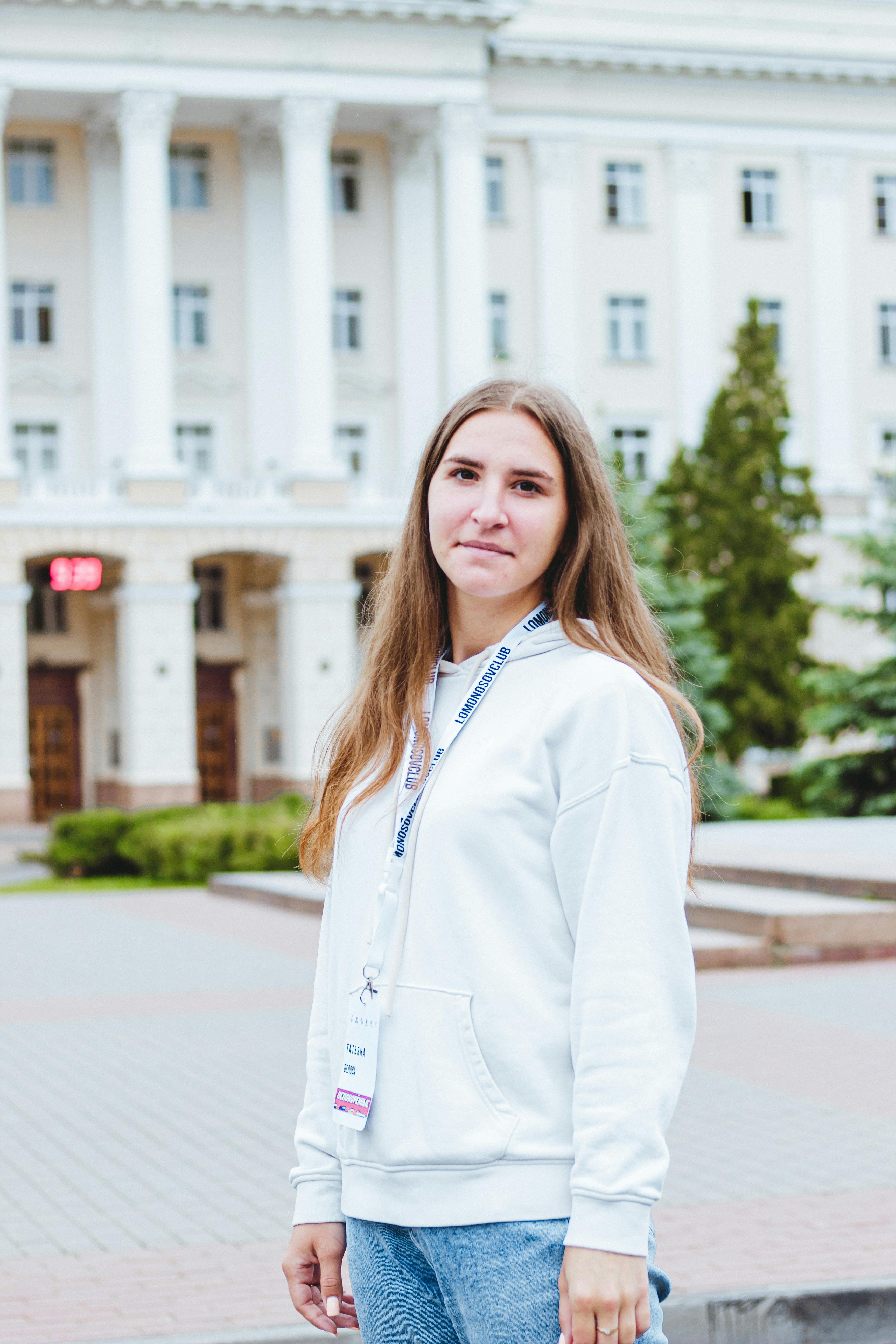 Young woman in a white hoodie smiling outside a college building.