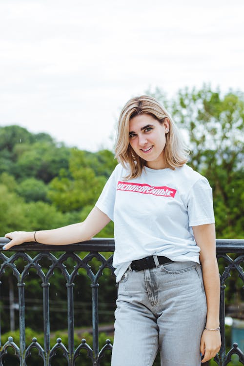 Woman in White Crew Neck T-shirt and Gray Denim Jeans Posing Beside the Metal Railing 