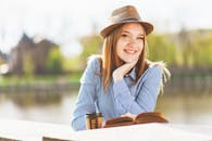 阳光明媚的日子里，一位戴着软毡帽的女人在户外微笑着看书、喝咖啡