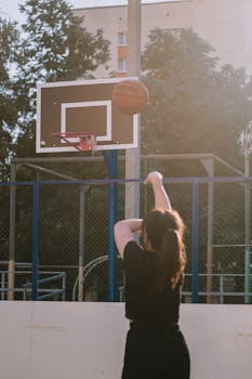 A woman shoots a basketball towards the hoop on an outdoor court during the day.