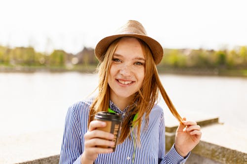 水域の近くに立っているプラスチック製のコーヒーカップを保持している白と青のストライプのボタンアップドレスシャツを着て笑顔の女性