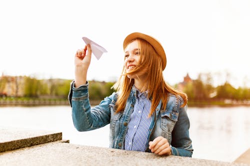 Woman in Blue Denim Button-up Jacket and Brown Hat About to Fly a Paper Plane Beside a Body of Water