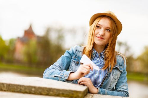 Free Smiling young woman wearing denim jacket and fedora, playfully holding a paper airplane outdoors. Stock Photo