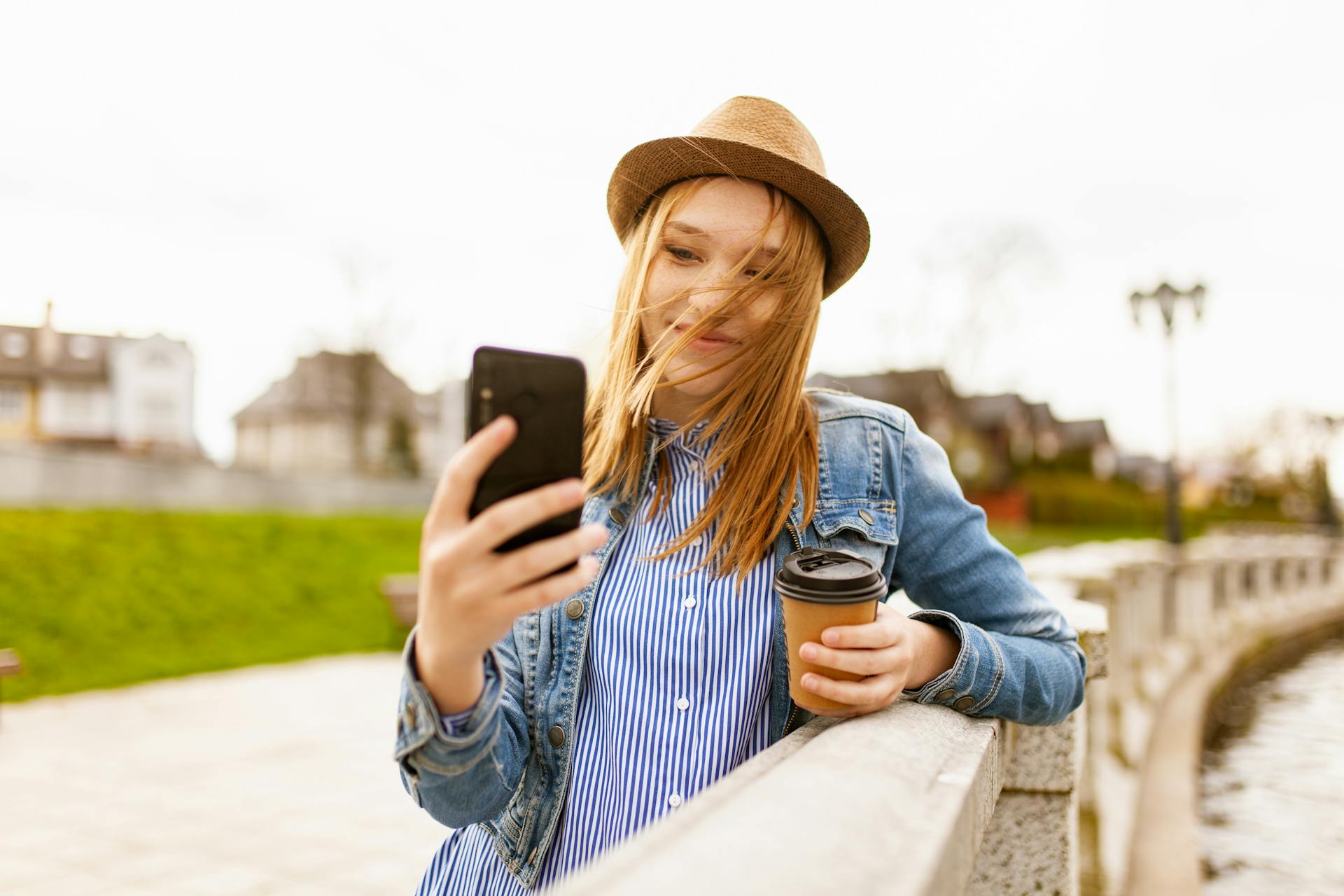 Smiling young woman with coffee outdoors using smartphone by a river.