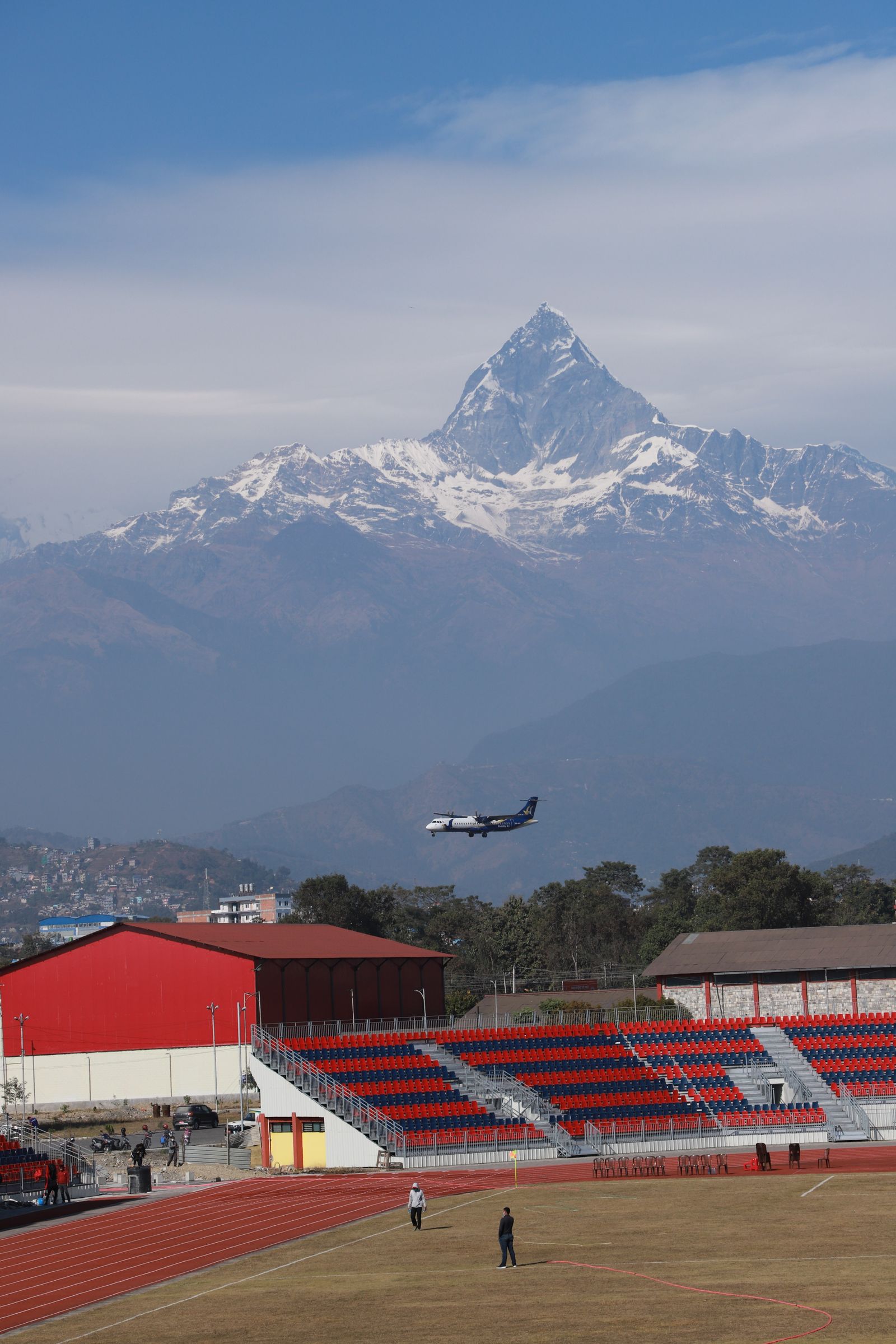 A turboprop descending into Pokhara beneath the snow pyramid of Machhapuchhre, a stadium's red stands in the foreground