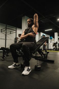 Man in black tank top stretching on a bench in a gym setting.