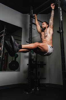 Athletic man with tattoos performing a workout on a pull-up bar in a gym.