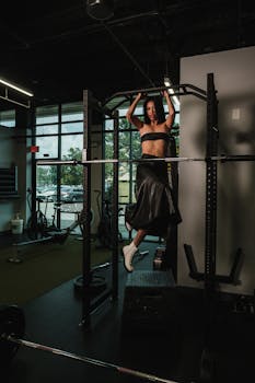 An adult woman in a gym performing a pull-up while wearing a stylish outfit.