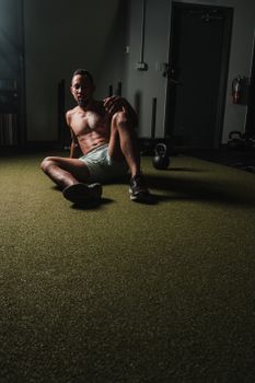 A muscular man is sitting shirtless on a gym floor, with dramatic lighting emphasizing his physique.