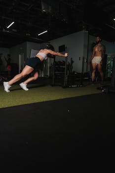 A muscular woman pushes workout equipment while a man observes in a gym setting.