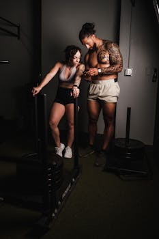 A tattooed couple stands in a gym, checking a smartphone during a fitness session.