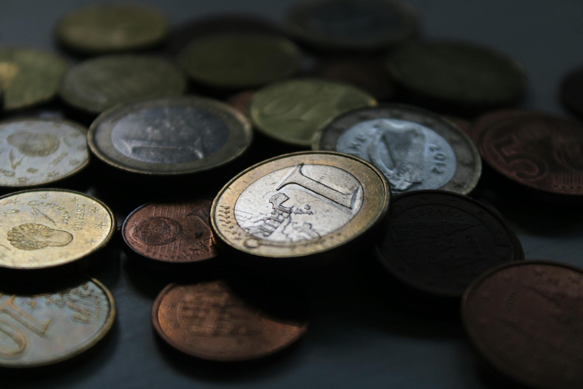 A close-up shot of Euro coins in various denominations scattered on a dark surface.