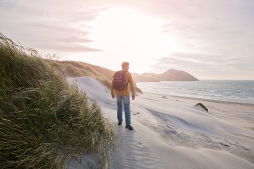 Free A man with a backpack walks along sandy dunes by the ocean at sunrise, enjoying nature. Stock Photo