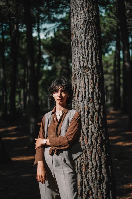 A woman leans against a tree trunk in a forest, dressed in stylish brown and gray attire.