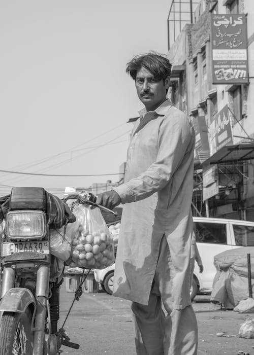 Man Standing by a Motorbike with a Bag of Fruit in Grayscale