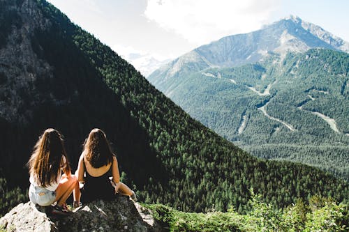Free Two Women Sitting on Rock Facing Mountains Under White Cloudy Skies Stock Photo