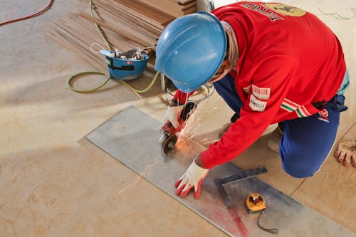 From above of faceless workman in uniform and protective hardhat using angle grinder while cutting metal layer on floor near tape measure at work