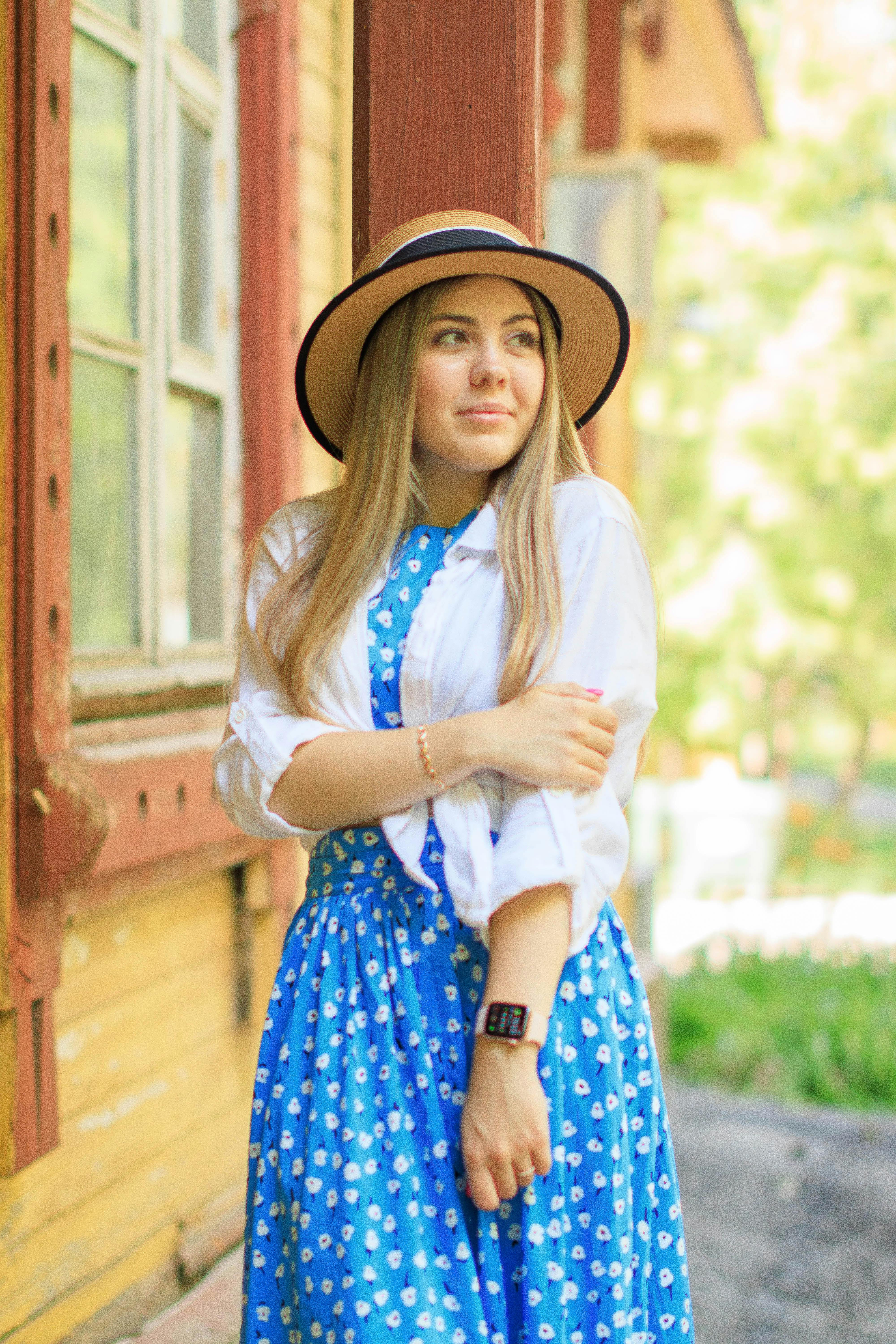 A young woman in a blue floral dress and hat stands by a rustic wooden building outdoors.