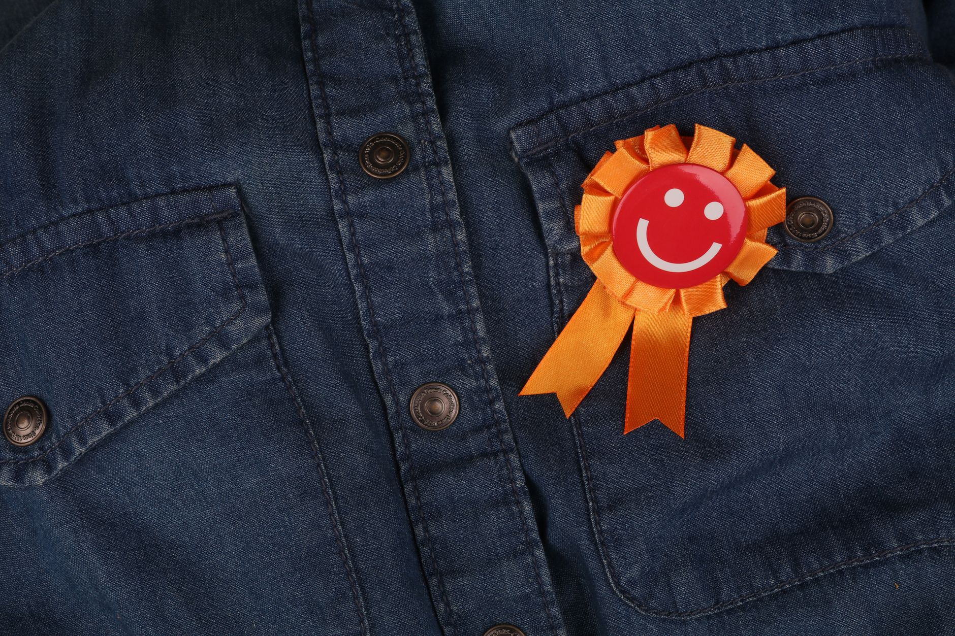 Blue denim shirt with orange ribbon pin featuring a smiley face, symbolizing happiness.