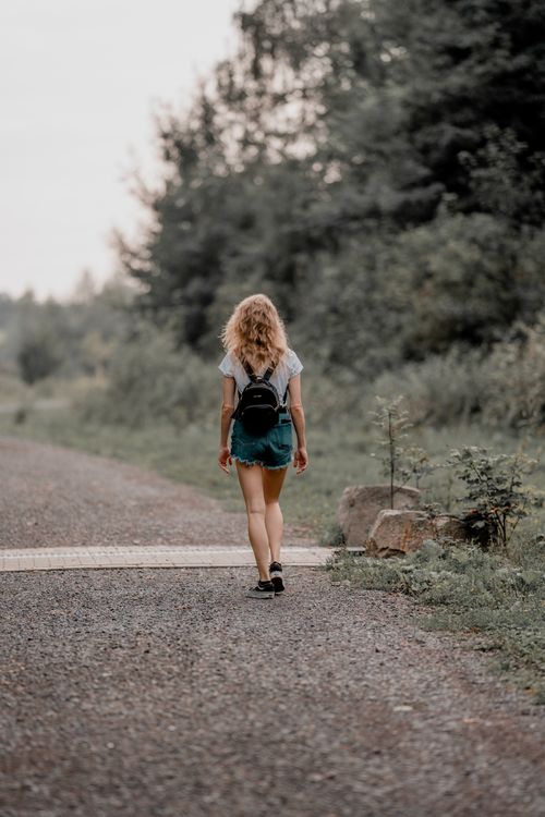 Free Back view of a young woman walking on a forest trail with a backpack, surrounded by nature. Stock Photo