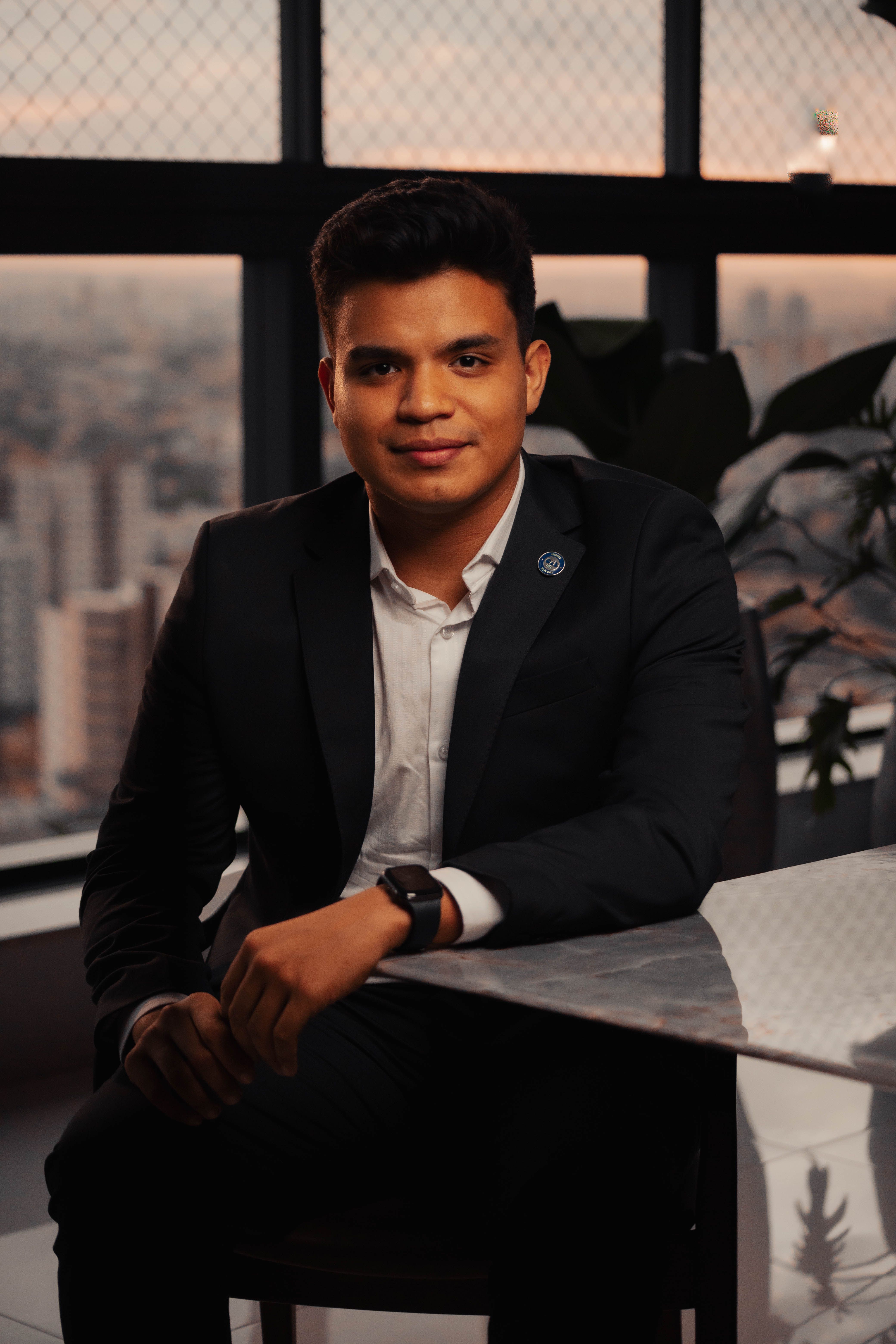 A young professional in a suit poses confidently in a modern office with a city view.