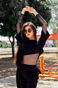 Trendy woman in sunglasses striking a pose with raised hands outdoors.