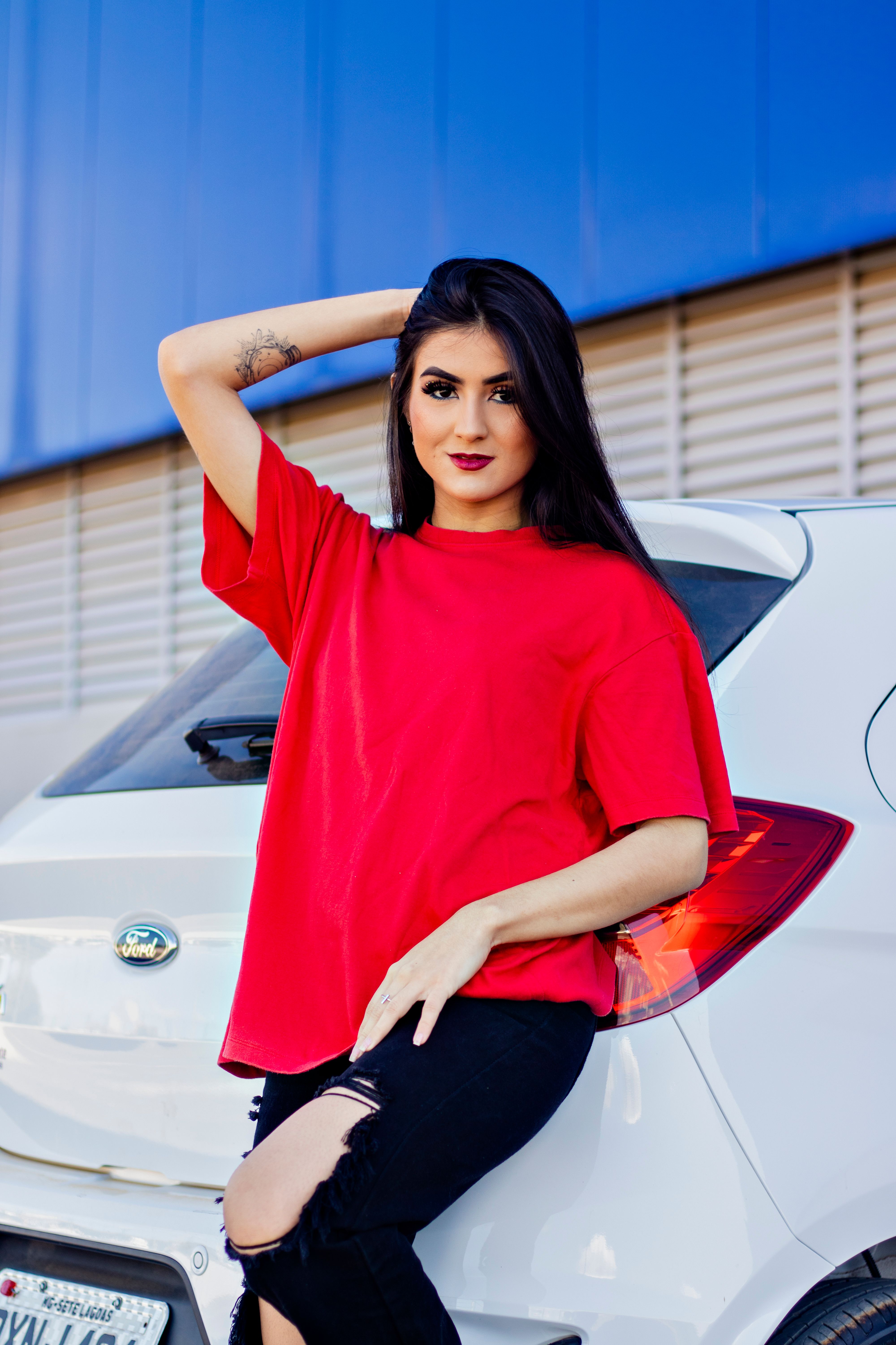 Woman in a red shirt leaning against a white hatchback, showcasing modern fashion.