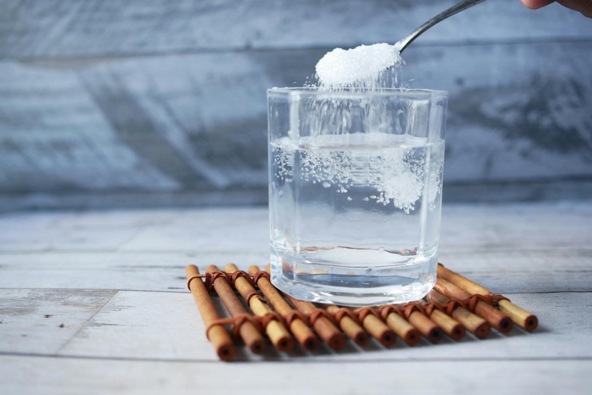 A spoonful of sugar being added to a glass of water on a wooden mat.