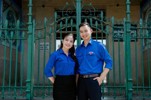 An Asian couple smiling in blue uniforms standing together outdoors in front of an ornate gate.