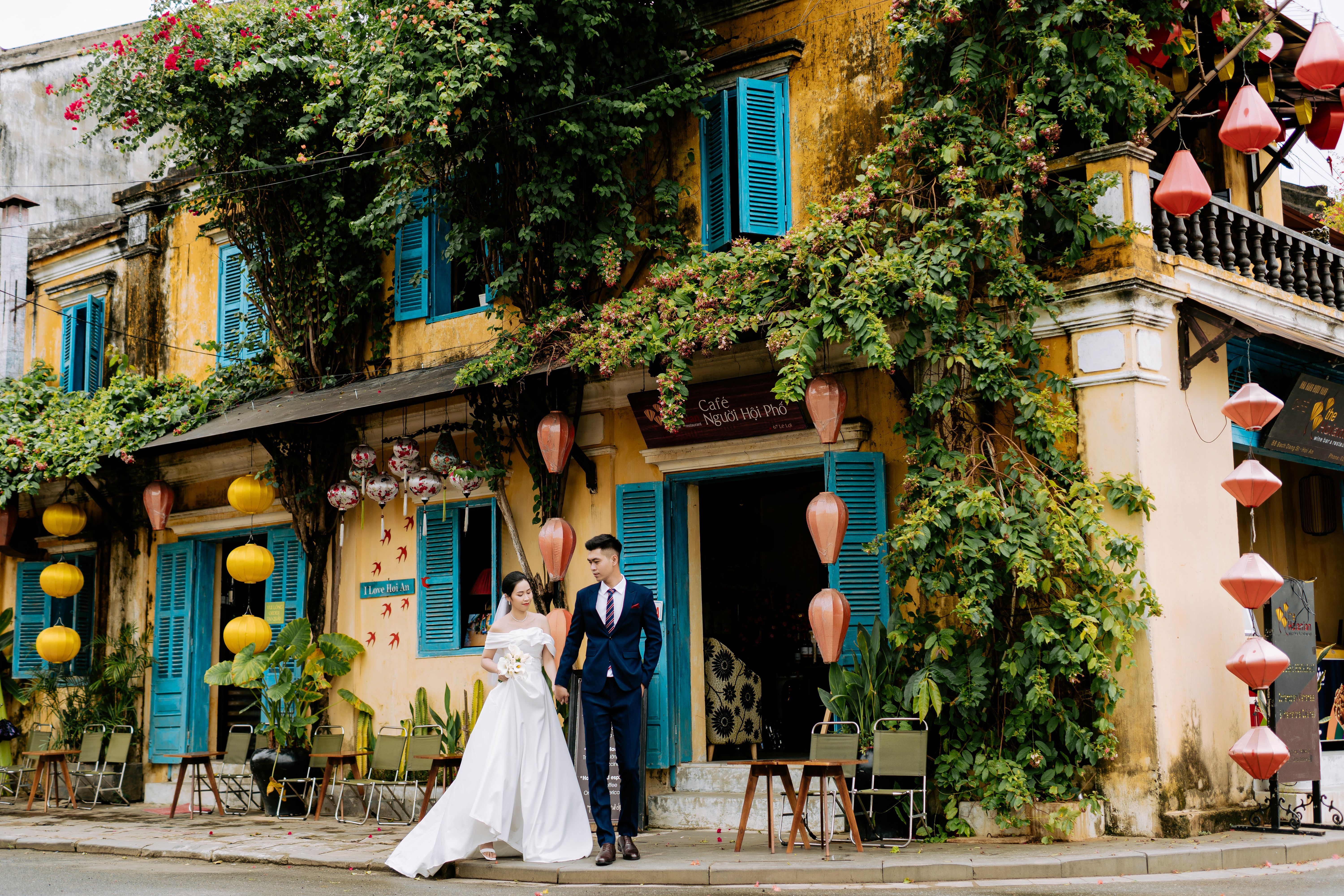 Newlyweds walking hand in hand past a charming café adorned with colorful lanterns and lush plants.