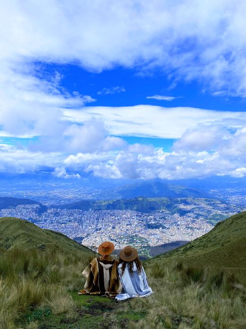 Free A couple in hats sits on a mountain peak, overlooking a sprawling city under a bright blue sky. Stock Photo