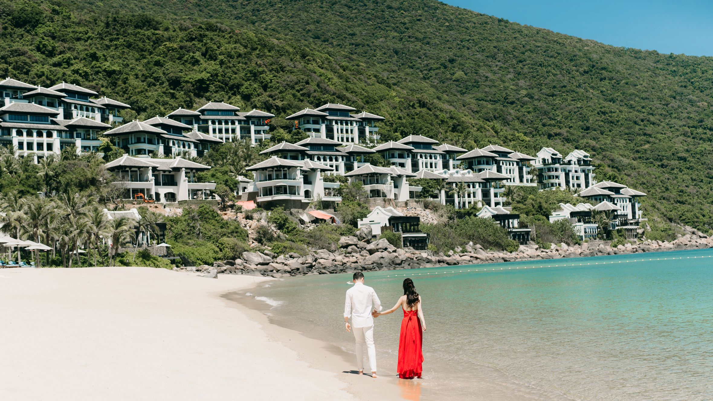 A couple walks hand in hand along a scenic beach next to luxurious villas, enjoying a sunny day.
