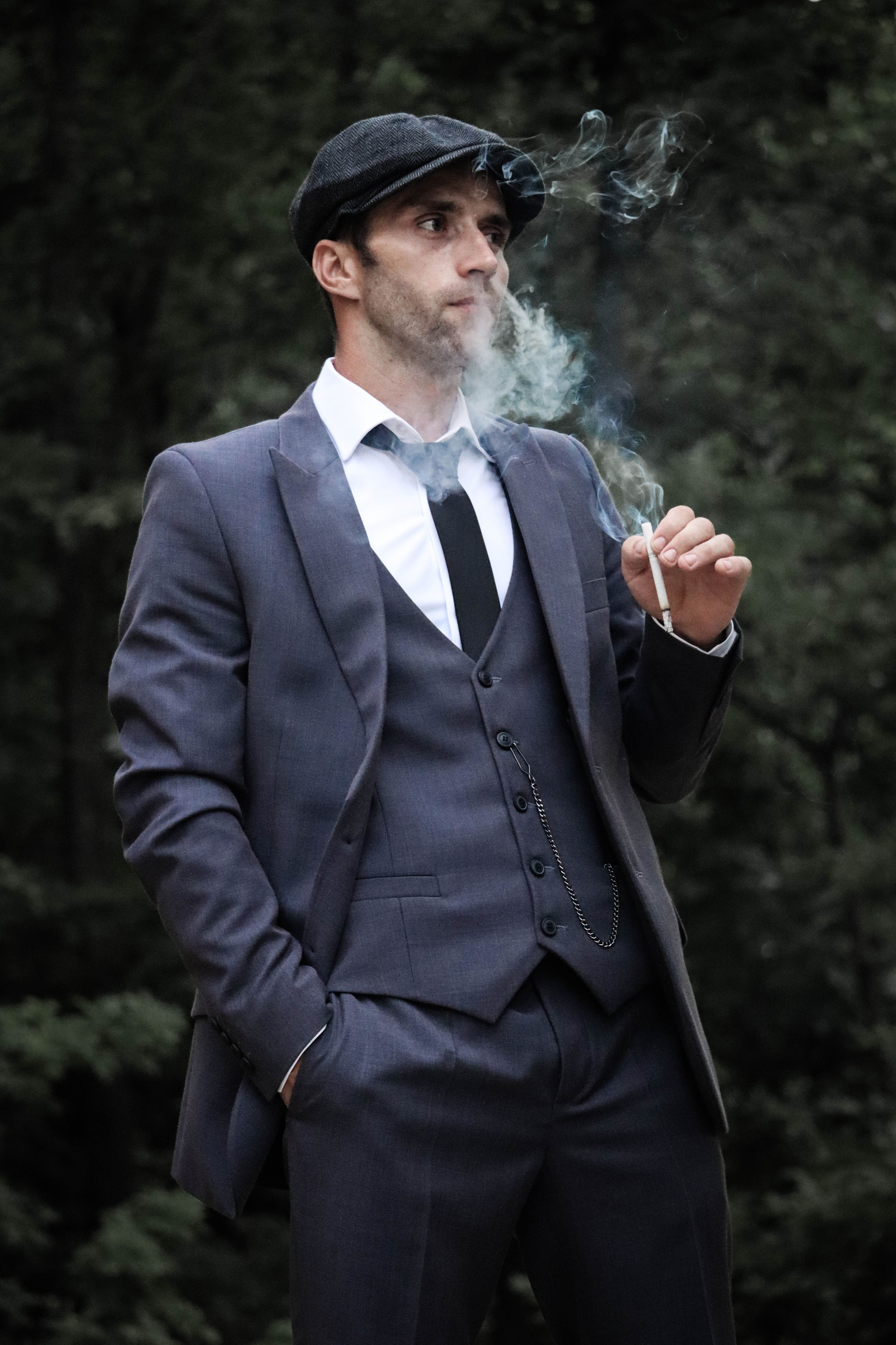 Stylish man in a gray suit and flat cap smoking outdoors, hand in pocket.