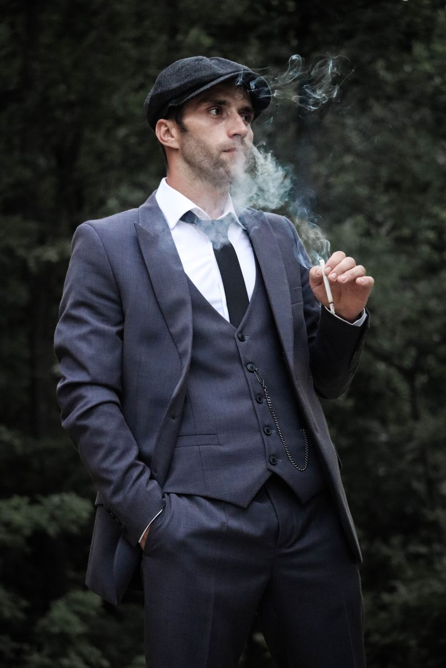 Stylish man in a gray suit and flat cap smoking outdoors, hand in pocket.
