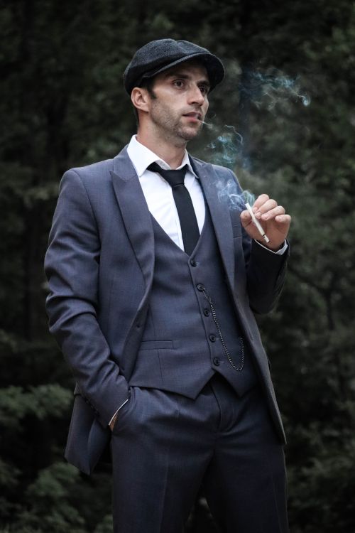 Free A fashionable man in a suit and flat cap smoking a cigarette outdoors, exuding style. Stock Photo