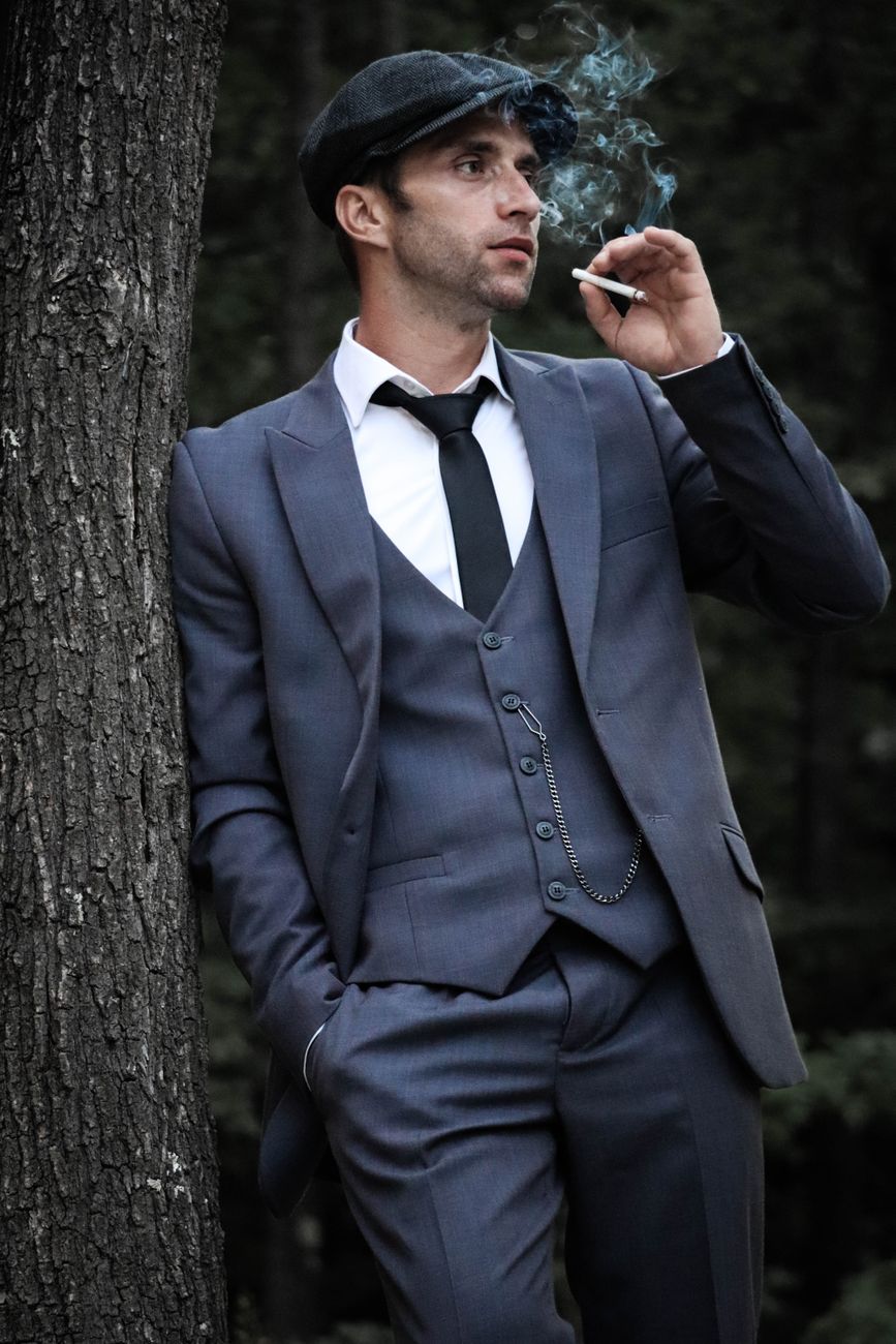 A stylish man in a formal suit and flat cap smoking outdoors, embodying classic fashion allure.
