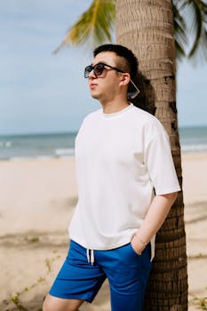 A man wearing sunglasses leans against a palm tree on a sunny beach, enjoying the summer day.